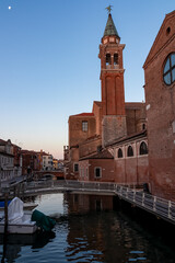 Fototapeta na wymiar Church of Saint James Apostle with view of canal Vena nestled in charming town of Chioggia, Venetian Lagoon, Veneto, Italy. Small boats floating in calm water creating romantic reflections. Tourism