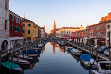 Church of Saint James Apostle with sunset view of canal Vena nestled in charming town of Chioggia, Venetian Lagoon, Veneto, Italy. Small boats floating in calm water creating romantic reflections