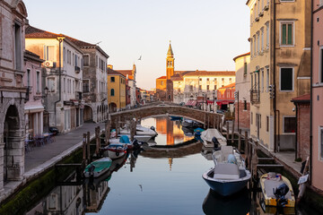 Church of Saint James Apostle with sunset view of canal Vena nestled in charming town of Chioggia, Venetian Lagoon, Veneto, Italy. Small boats floating in calm water creating romantic reflections