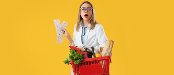 Shocked young woman with shopping basket of food and receipts on yellow background. Price rise...