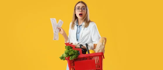 Poster Shocked young woman with shopping basket of food and receipts on yellow background. Price rise concept © Pixel-Shot