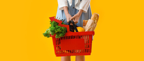 Young woman with shopping basket of food on yellow background, closeup. Price rise concept