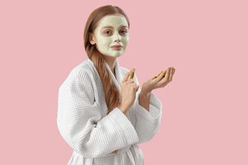 Young woman in bathrobe with applied turmeric mask and roots on pink background