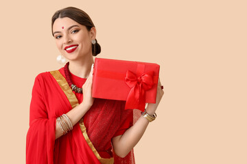 Beautiful young Indian woman in sari with gift box on beige background