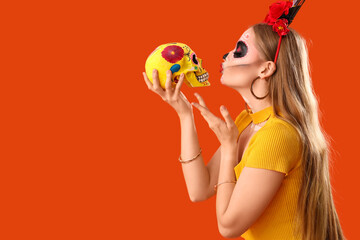 Young woman with painted skull blowing kiss on orange background. Celebration of Mexico's Day of...