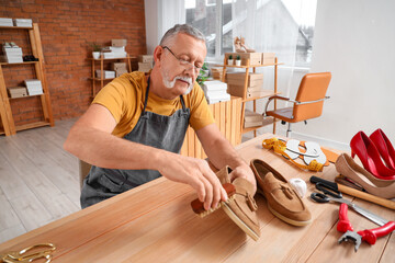 Mature shoemaker brushing shoe at table in workshop - obrazy, fototapety, plakaty