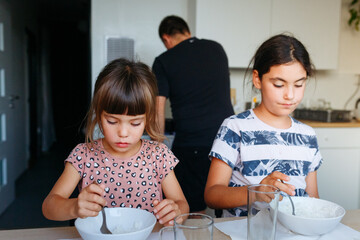 two siblings eating lunch at home at white table with father cooking in background