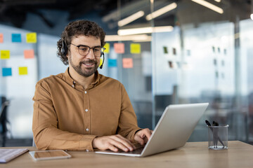 Cheerful male professional using laptop and wearing a headset in a modern office environment, displaying positivity and productivity.