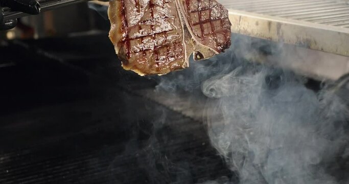 T-bone meat steak on barbecue charcoal grill. Chef takes ready steak off the grill. Grilling BBQ meat. Smoke rising over juicy beef steak
