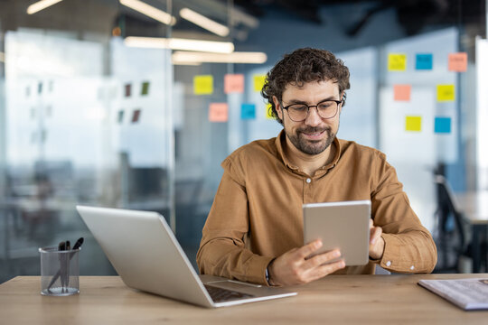 Casually dressed businessman working with a digital tablet and laptop at a wooden desk in a contemporary office setting. Productive and relaxed atmosphere.