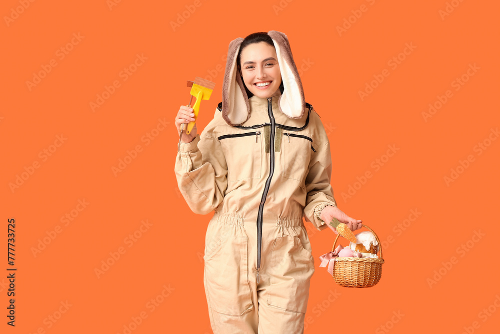 Poster Happy female beekeeper in bunny ears with Easter basket and tools on orange background