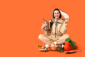 Happy female beekeeper in bunny ears with Easter baskets, toy carrots and tools sitting on orange...