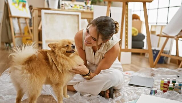 Young hispanic woman artist sitting on floor, smiling lovingly and kissing her adorable pet dog in a colourful art studio