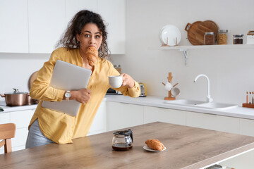 Hurrying young African-American woman with croissant, coffee cup and laptop in kitchen