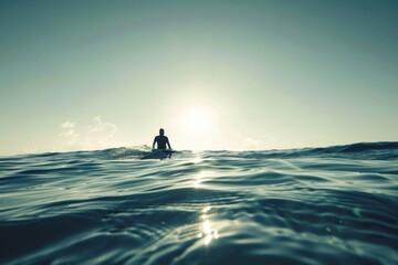 Surface of the ocean with clear sky and sun overhead and silhouette of a surfer on the horizon