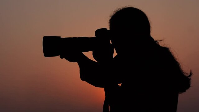A young photographer is taking pictures with a professional camera in the color of a golden summer sunset sky.