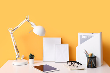 Comfortable workplace with tablet computer, desk lamp and notebooks near orange wall