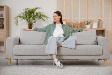 Pretty young woman sitting on grey sofa in living room - obrazy, fototapety, plakaty