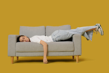 Tired young woman lying on grey sofa against yellow background - obrazy, fototapety, plakaty