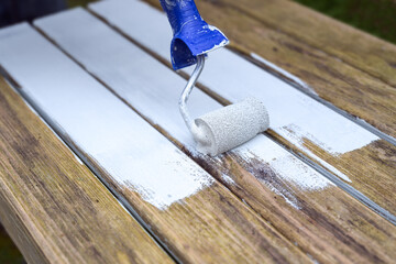 Painting a weathered wooden garden table with a paint roller and white wood protection coat, restorating old furniture for the outdoor season in spring, summer and autumn, copy space - 777745326