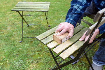 Man is cleaning and restoring wooden outdoor furniture, sanding the weathered wood to remove algae before oiling or painting for a neat garden season, copy space