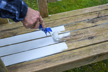 Man is painting a weathered wooden outdoor bench with a white weather protection paint using a lacquer roller, refreshing old outdoor furniture for the garden season, copy space - 777745756