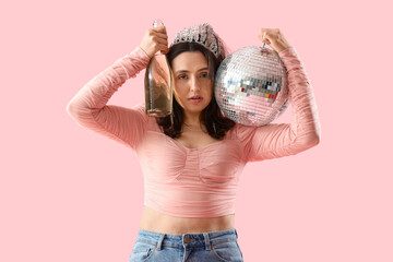 Young woman with hangover and disco ball putting bottle to her head after Birthday party on pink...