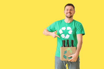 Happy young man in t-shirt with recycling logo pointing at cardboard box with glass bottles on...