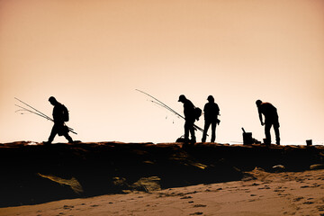 Silhouette of 4 fishermen along the stone breakwater at the inlet at Ocean City MD. 2 men with fishing poles and gear head for their favorite fishing spot along the rocks. Atlantic Ocean