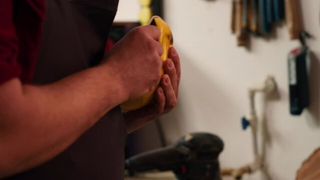 Carpenter putting sandpaper on piece of plastic, creating abrasive sponge to smooth surfaces. Man preparing necessary gear for furniture assembling job in studio, close up shot, camera A