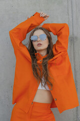 young girl in sunglasses and orange pantsuit at concrete wall
