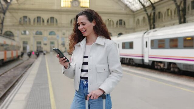 Happy young woman of young traveling woman with luggage using phone at platform station while waiting the train. Technology and travel concept.
