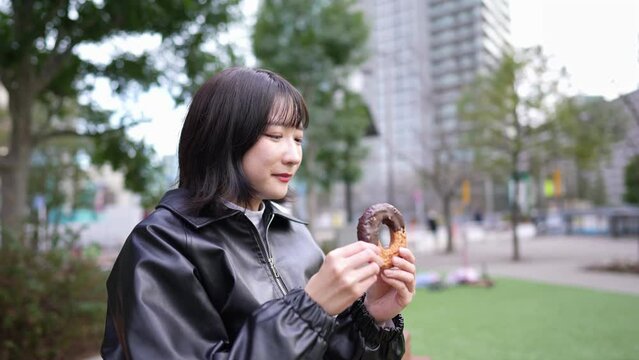 Slow motion video of a Japanese woman in her 20s eating donuts in a park around Gotanda Station in Shinagawa-ku, Tokyo in winter 冬の東京都品川区五反田の駅周辺の公園でドーナツを食べる20代の日本人女性のスローモーション映像