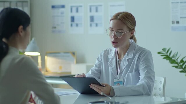 Female doctor taking clipboard with medical records from patient, reading them and asking questions about health during appointment in clinic