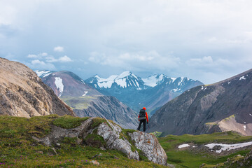 Man in vivid red jacket admire alpine scenery on green grassy hill among rocks near precipice edge. Guy with backpack on pass enjoying few big snowy pointy peaks. Three large snow peaked tops far away - 777819135