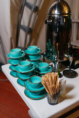 Elegant Coffee Service with Turquoise Cups and Saucers. A coffee serving station featuring shiny metal coffee urn, turquoise ceramic cups with saucers, and wooden stir sticks, arranged neatly for a ga