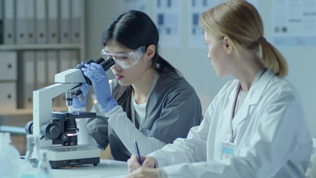 Young Asian laboratory technician using microscope and telling results on medical research to Caucasian female doctor taking notes on paper when working together in clinic