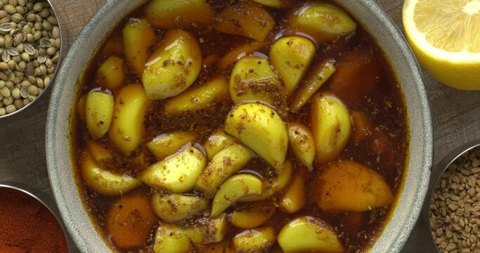 Top view of garlic pickle, spicy, tangy, and mouth-watering side dish from India. Around the bowl with pickled garlic are containers with spices used for pickling. Zoom in while rotating.