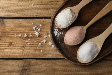 Different natural salt in spoons on wooden table, flat lay. Space for text