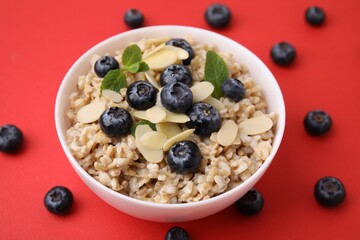 Tasty oatmeal with blueberries, mint and almond petals in bowl surrounded by fresh berries on red...