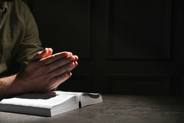 Religion. Christian man praying over Bible at table against black background, closeup. Space for...