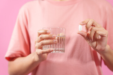 Graciously, woman hand holds  bottle of cod liver oil and a glass of water against a pink backdrop, illustrating health, hydration, and nutritional supplementation.