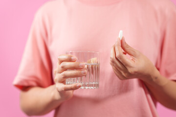 Graciously, woman hand holds  bottle of cod liver oil and a glass of water against a pink backdrop,...