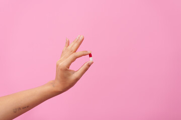 A female hand delicately holds a white pill between fingers against a pink isolated background,...