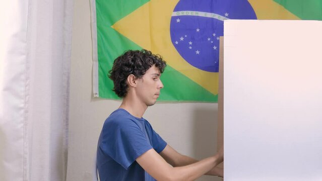 Jovem homem brasileiro escolhendo o seu candidato na urna eletrônica. Na parede, a bandeira do Brasil decora o local de votação. A imagem retrata as eleições brasileiras