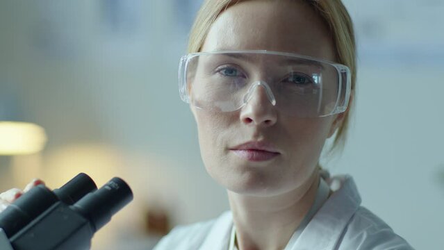 Female chemist in protective glasses looking through microscope and posing at camera at workplace in medical laboratory. Video portrait