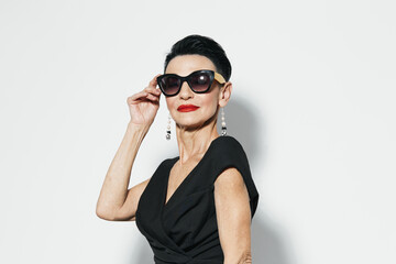 Elegant senior woman in black dress and sunglasses posing in front of white wall for a fashion...