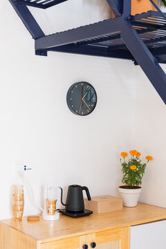 A wooden shelf with glasses and electric kettle under a wall clock