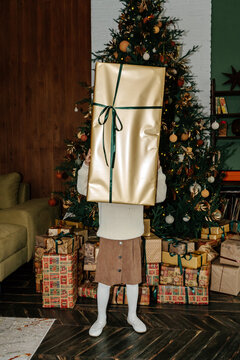 Little girl in skirt covering face with big gift near Christmas tree