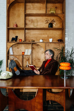 Mature man reading book with feet on table in stylish office